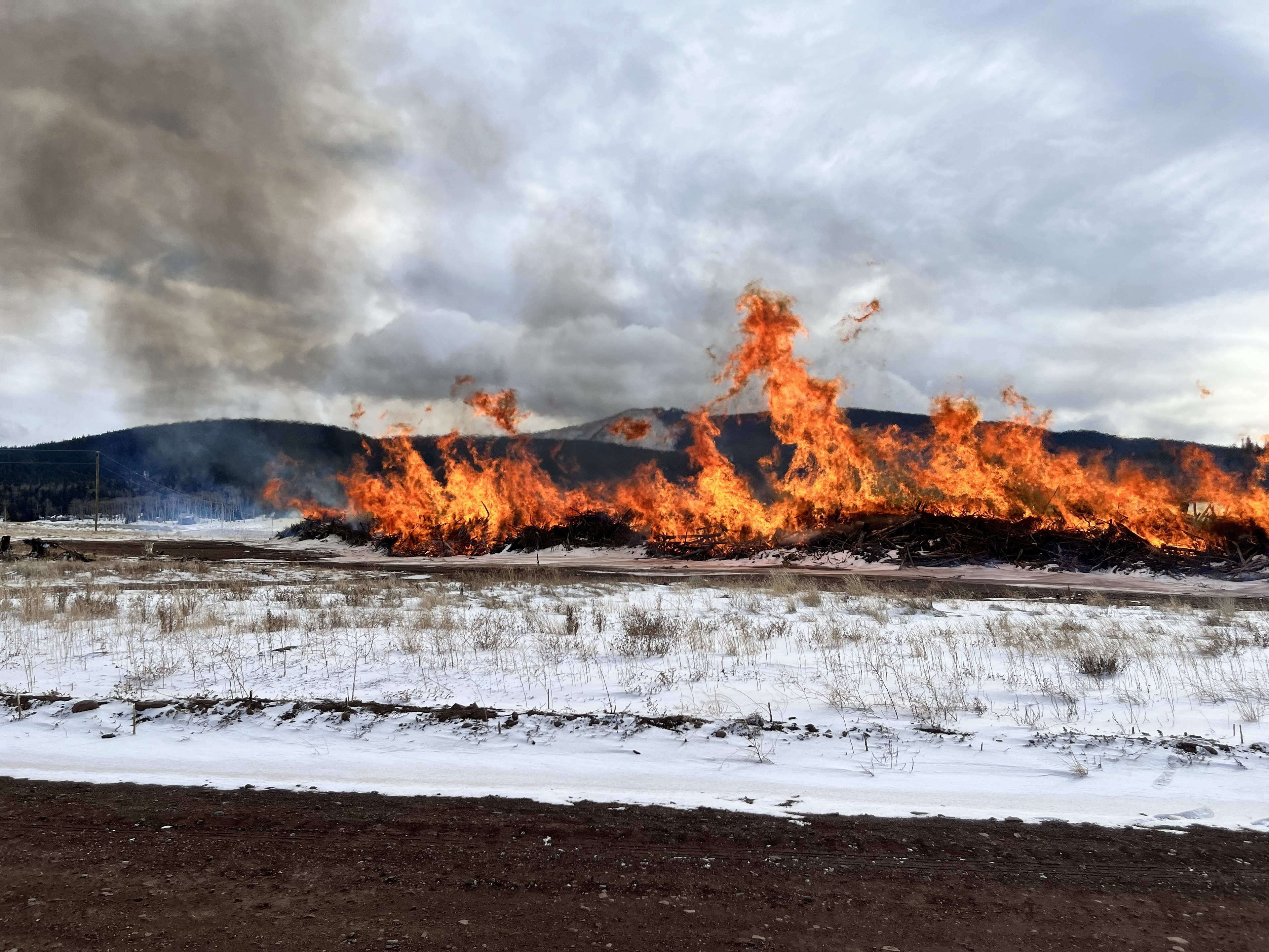 Large controlled slash pile burn in a snowy field with mountains in the background